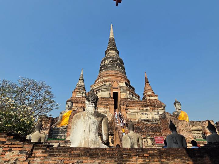 Wide-angle view of a grand Ayutthaya chedi flanked by Buddha images with visitors climbing central stairs under clear blue sky