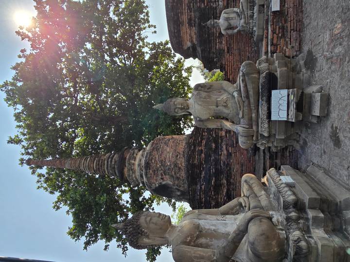 Weathered Buddha statues beneath a tall brick pagoda and leafy tree with sun flare in the corner