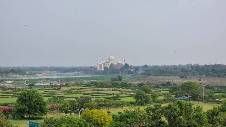 Distant view of the Taj Mahal across a broad river plain surrounded by green gardens and trees under hazy sky