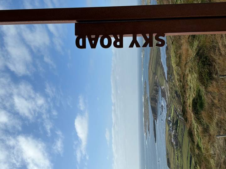 Scenic coastal panorama from a hilltop viewpoint with an iron sign reading SKY ROAD overlooking ocean inlets and islands under a blue sky with scattered clouds