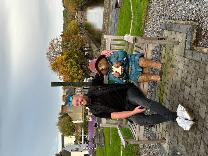 Man sitting on a bench next to a Paddington Bear statue beside a tranquil canal lined with autumn trees