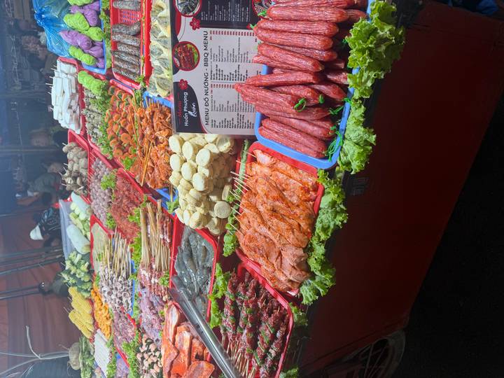 Colorful street-market barbecue stall displaying skewers, sausages and fresh produce under bright lights