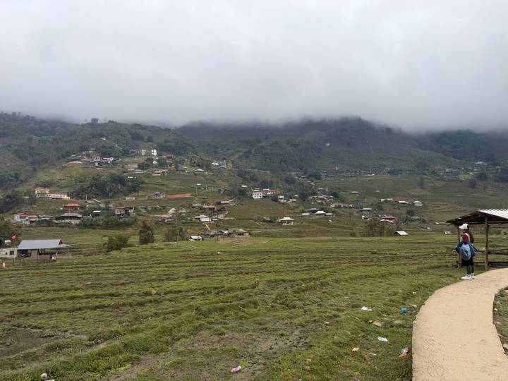 Mist-covered valley with scattered houses and terraced fields viewed from a pathway where a traveller pauses with a backpack