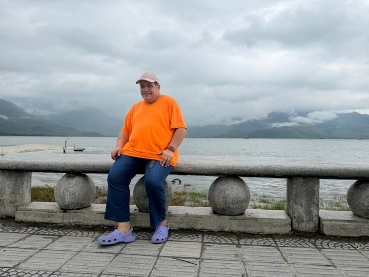 Traveller in bright orange shirt sits on a stone balustrade beside a vast lake with cloudy mountains beyond