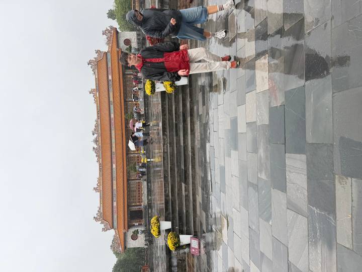 Visitors with umbrellas walk across rain-soaked marble courtyard toward a traditional Vietnamese palace building