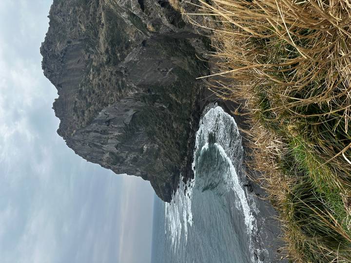 Dramatic seaside cliff on a cloudy day with waves crashing against a dark sand beach on Jeju Island.