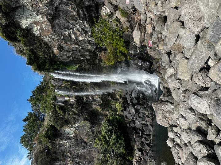 Twin-stream waterfall plunging down a rocky cliff into a pool surrounded by boulders and lush greenery.