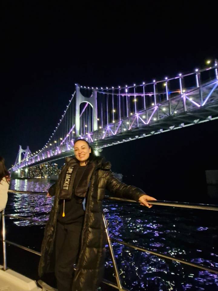 Smiling traveler on a night boat cruise with Busan’s illuminated Gwangan Bridge glowing purple behind.