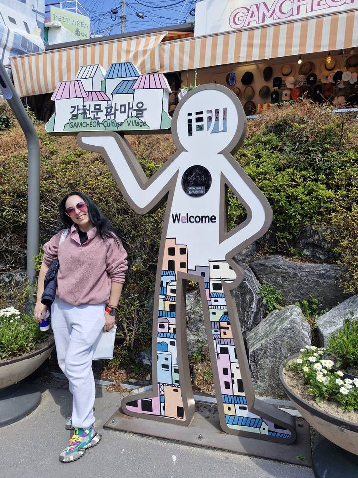 Traveler posing beside a playful welcome sign figure in a leafy outdoor setting.