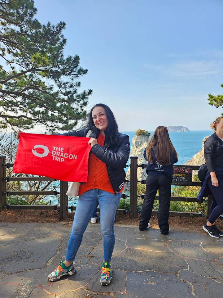 Woman proudly holding a red travel flag at a coastal viewpoint with sea stacks and blue water behind.