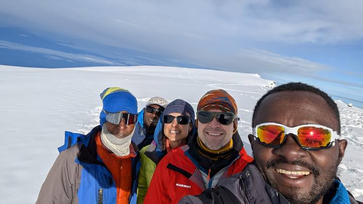 Five climbers wearing goggles smile in a selfie on a broad, snow-covered summit plateau under clear skies.