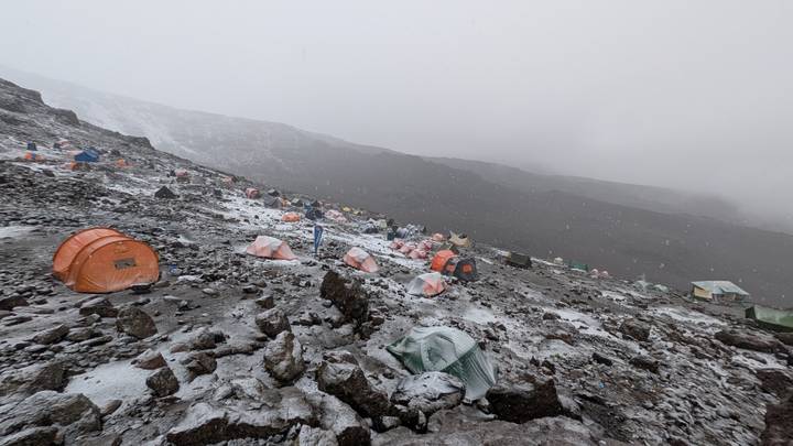 A high-altitude campsite with dozens of orange tents scattered over a rocky, lightly snow-dusted slope in misty weather.