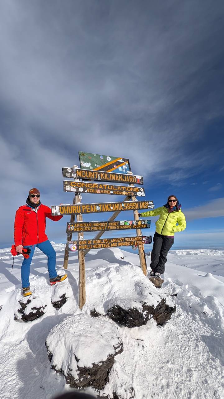 Two climbers stand next to the colourful wooden Uhuru Peak sign on Mount Kilimanjaro under a clear blue sky.