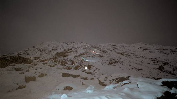 Nighttime scene of snow-covered slope with small lines of climber headlamps ascending in darkness.