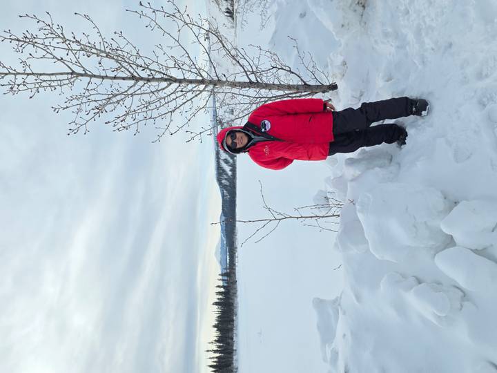 Explorer in a red parka stands beside a frozen lake and snowy forest mountains.