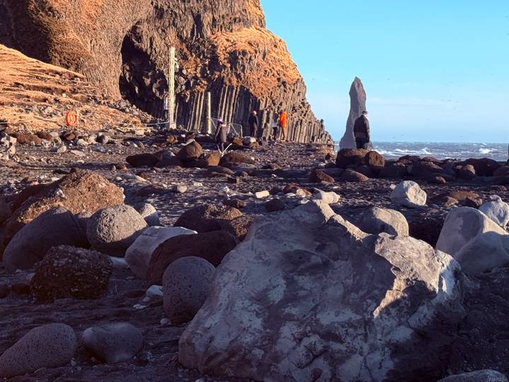 Visitors explore basalt column cliffs on a black sand beach beside roaring waves