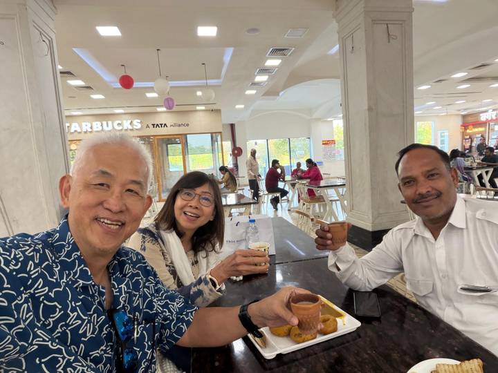 Three travelers smile while enjoying drinks at a food court table inside a shopping mall.