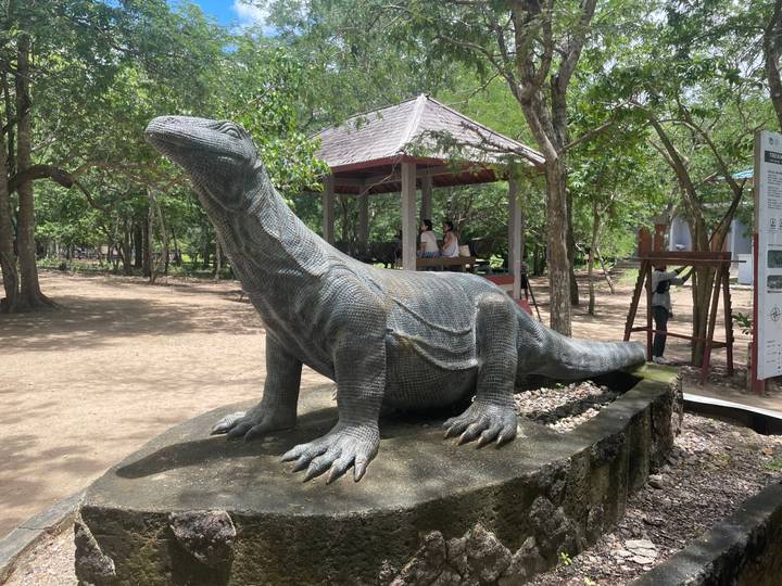 Life-size Komodo dragon statue on a stone plinth with visitors resting in a shaded pavilion behind.