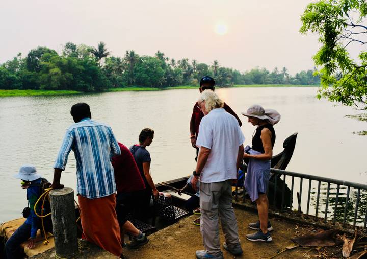 Travelers board a small wooden ferry on a tranquil river as the sun sets through haze.