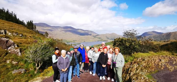 Tour group posing on a scenic overlook above a lake and mountains in Ireland under partly cloudy sky.