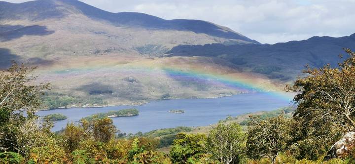 Bright rainbow arcing over a tranquil Irish lake with rugged mountains and islands.