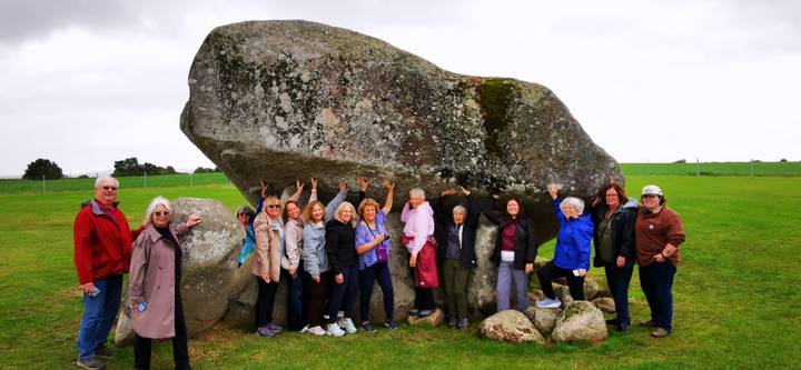 Large group standing under and supporting a massive glacial erratic boulder in a green Irish field.