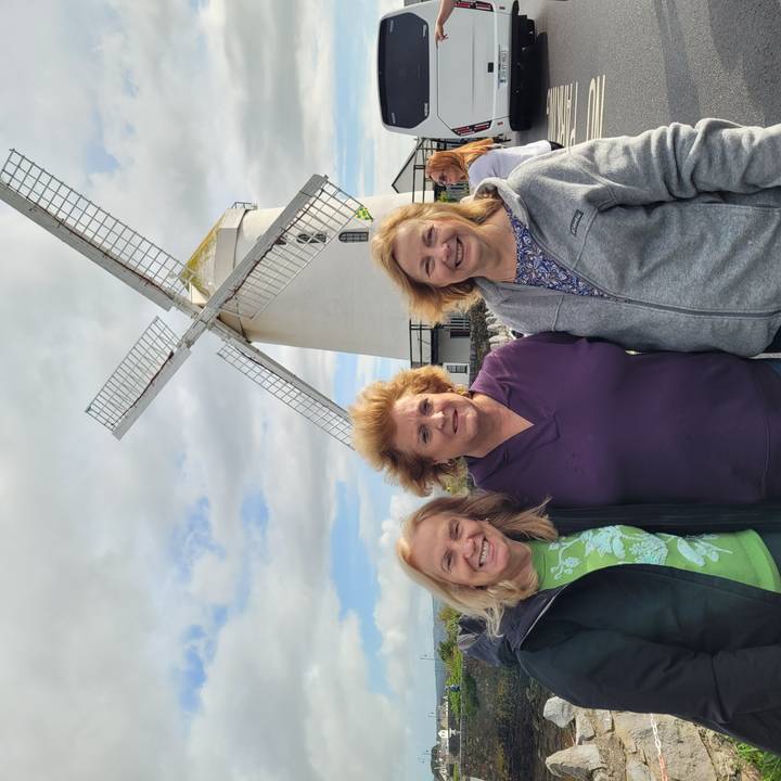 Three women smiling in front of a white windmill with cross blades under partly cloudy sky.