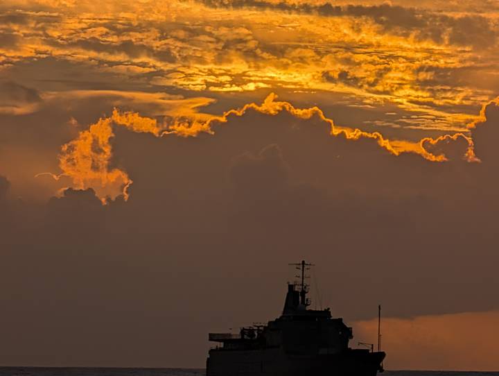 Dramatic orange clouds and silhouetted ship mast dominate a fiery evening sky.