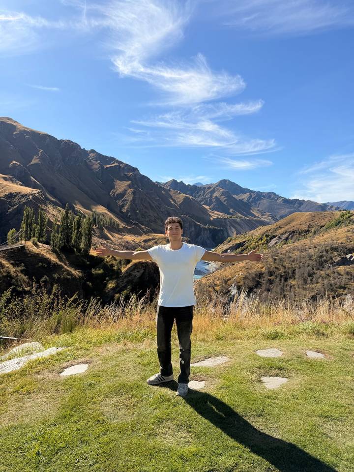 A young man in a white T-shirt spreads his arms wide before a dramatic New Zealand river gorge and mountains.