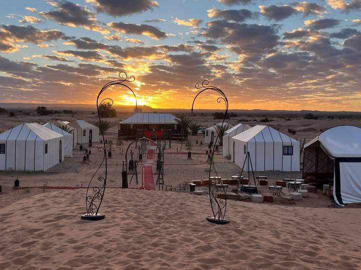 Luxury white desert tents and ornate iron arches glow under a vivid orange sunset in the Sahara dunes.