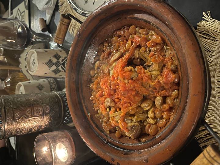 A bubbling Moroccan tagine filled with meat, tomatoes and beans sits on a clay dish at dinner.
