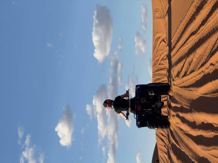 A rider on a blue quad bike carves tracks across golden desert dunes beneath a deep blue sky.