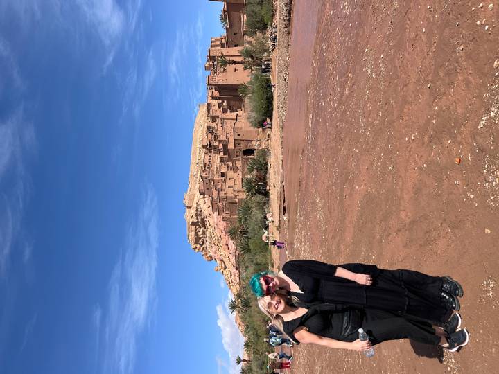 Two women stand smiling before the earthen kasbah of Aït Ben Haddou under a blue sky.