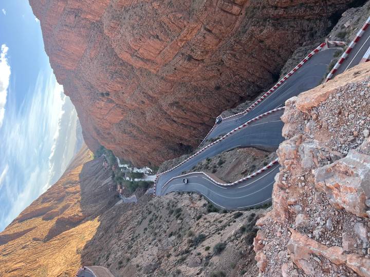 A dramatic serpentine road winds through steep red cliffs of the Dades Gorge with tiny cars below.