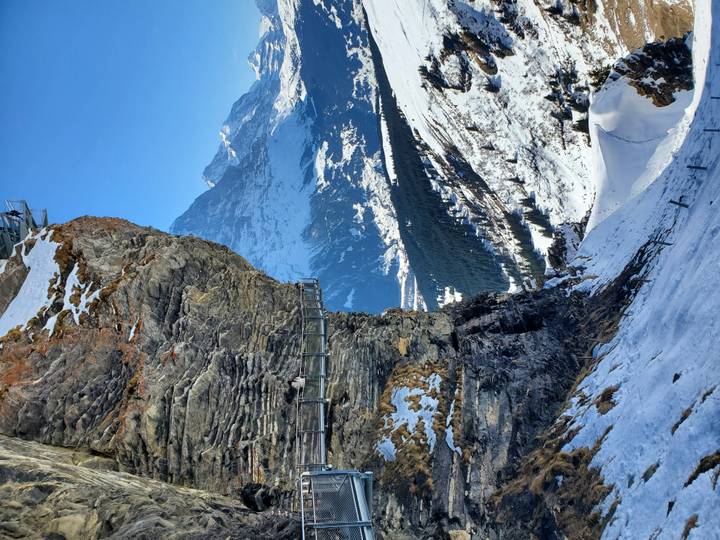 A cliff-edge metal walkway clings dramatically to a snowy alpine rock face with high peaks beyond.
