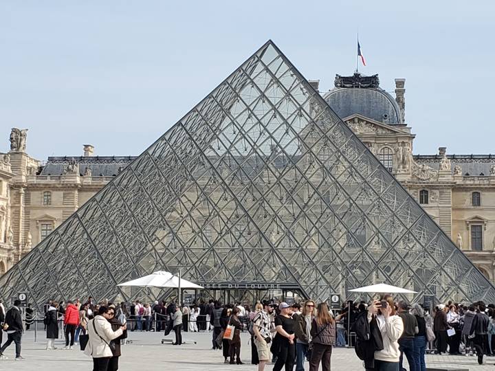 Crowds gather outside the Louvre Glass Pyramid on a clear day in Paris.