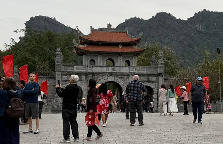 Visitors with red flags gather before an ornate stone gate backed by limestone peaks.