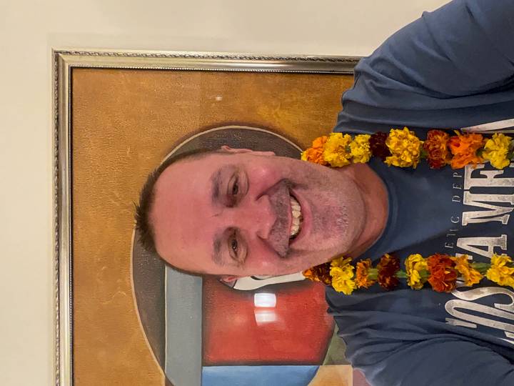 Smiling man wearing a marigold flower garland taking an indoor selfie against a framed artwork backdrop.