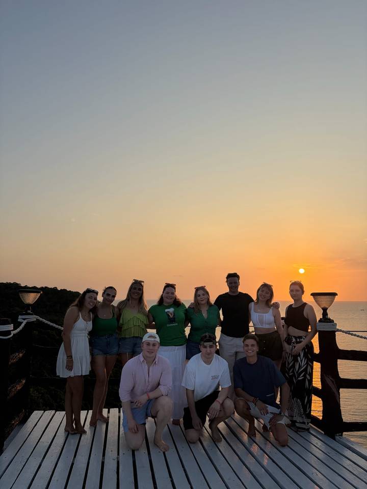Silhouetted friends stand together on a deck at sunset with orange sky over the sea.