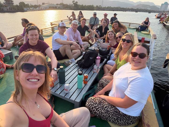 Group of travellers smiling on a wooden boat deck enjoying drinks during golden evening light.