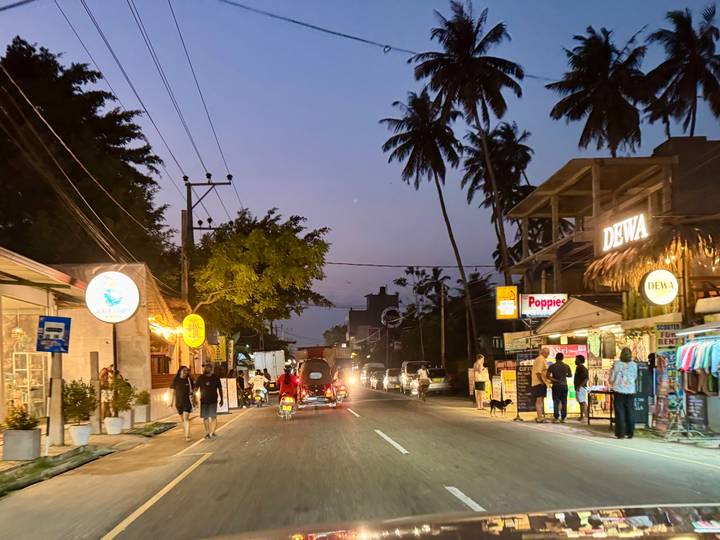 Dusk street scene lined with small shops, tuk-tuks, pedestrians and palm silhouettes in a Sri Lankan coastal town.