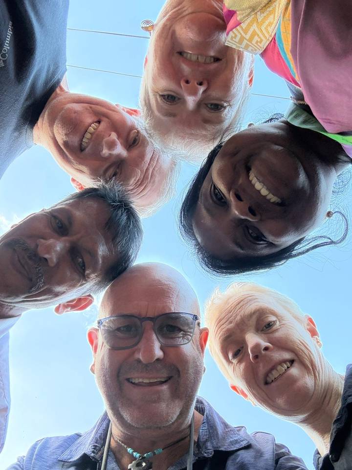 Playful selfie shot of five travellers forming a circle and looking down at the camera against blue sky.