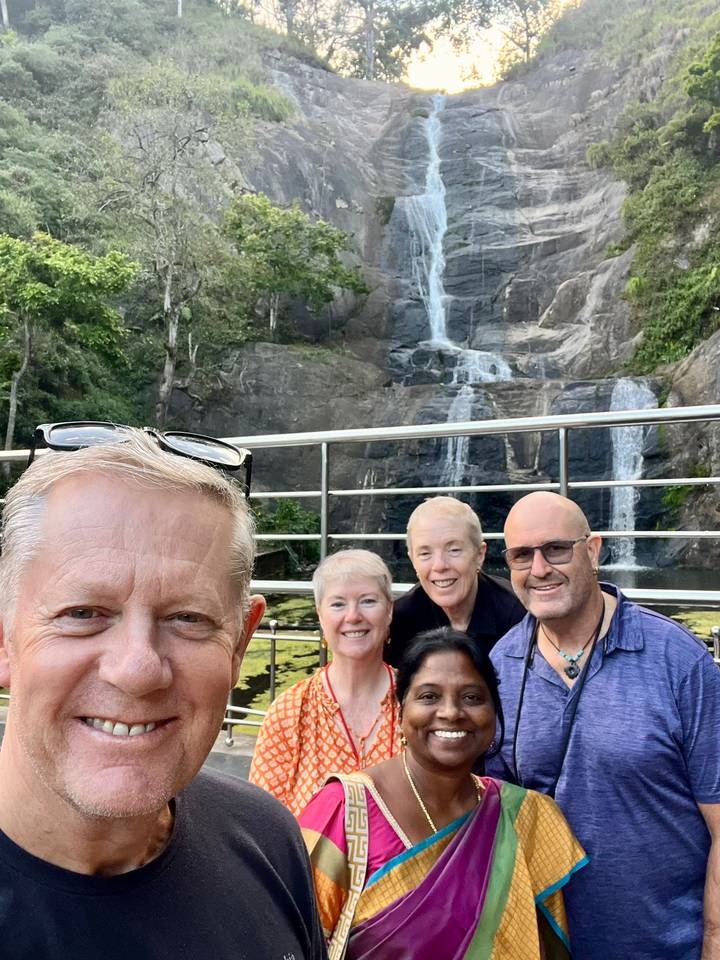 Group selfie in front of a tiered waterfall framed by forest and metal railing.