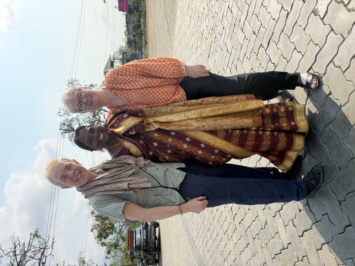 Two foreign travellers and their local guide in sari smile on a paved courtyard under hazy sun.