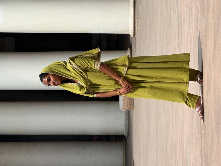 Graceful young woman in olive-green traditional dress stands poised between white columns.
