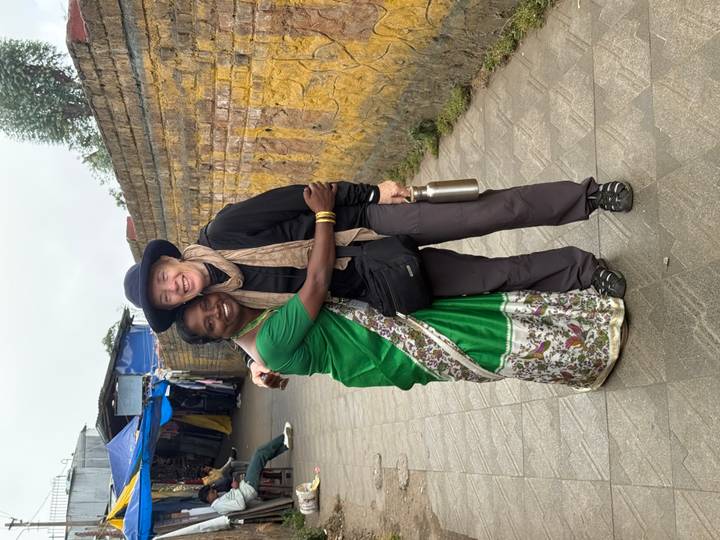 Two women share a warm hug on a stone walkway lined by colourful market stalls and yellow wall.