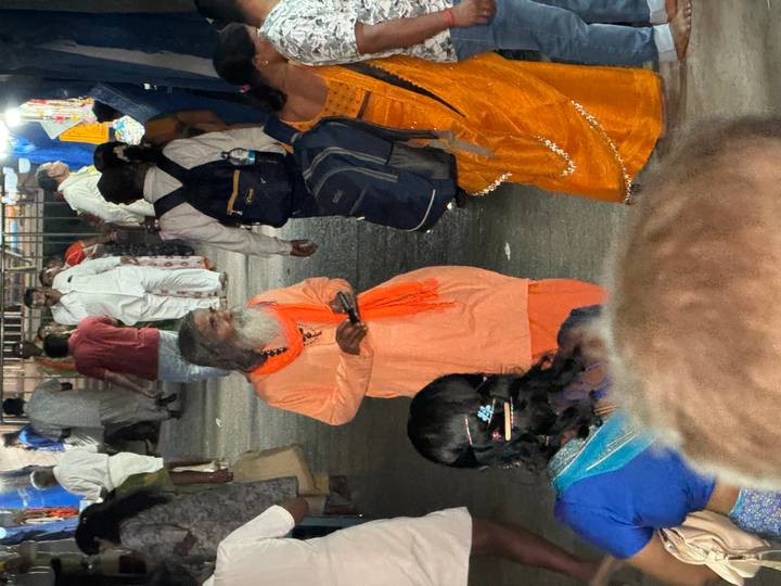 Bearded sadhu in orange robes stands amid evening market crowd lit by artificial lights.