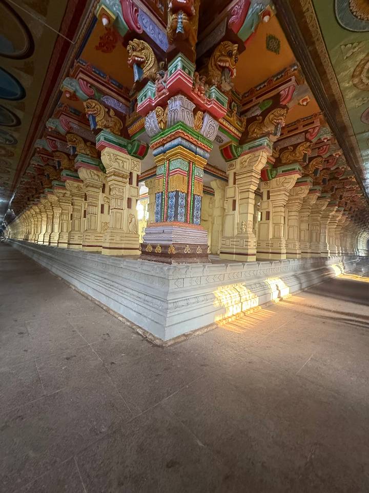 Grand ornate corridor of a South Indian temple with richly painted columns and slanting sunlight illuminating stone floors