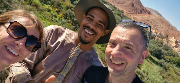 Casual selfie of three smiling travellers with green valley and red Atlas mountains behind