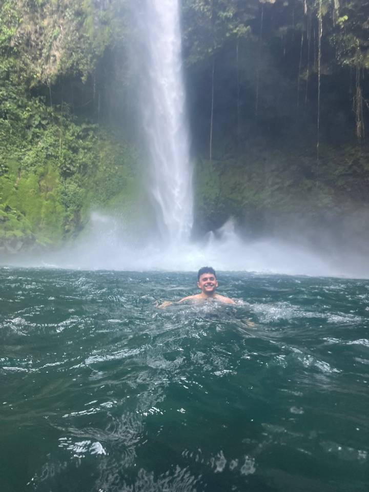 Young man swimming in a turquoise pool beneath a powerful jungle waterfall, smiling toward camera